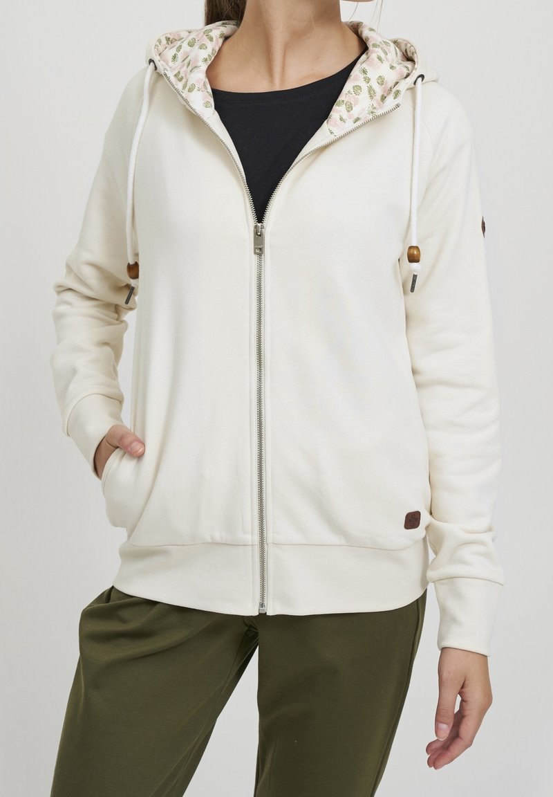 Sudadera con cremallera color crema, con un forro de capucha estampado, que cuenta con cordones, bolsillos laterales y un parche con logo en la parte frontal.