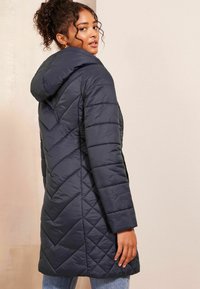 Manteau matelassé bleu marine avec capuche, présentant une couture en chevron, des manches longues et une coupe ajustée, exposé en vue arrière trois-quarts.