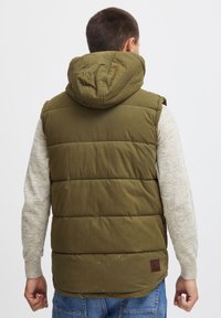 Gilet rembourré vert avec capuche, présentant une surface texturée, un design cousu et une écusson brun au dos, porté sur un pull léger.