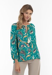 Blusa turchese con motivo paisley in arancio, giallo e viola. Presenta un collo con cordoncino e nappine e maniche lunghe arricciate.