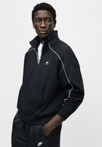 Μαύρο φούτερ με φερμουάρ από μαλακό ύφασμα, με λευκή διακόσμηση, τσέπη κάγκουρο και μικρή ετικέτα με λογότυπο στο στήθος.