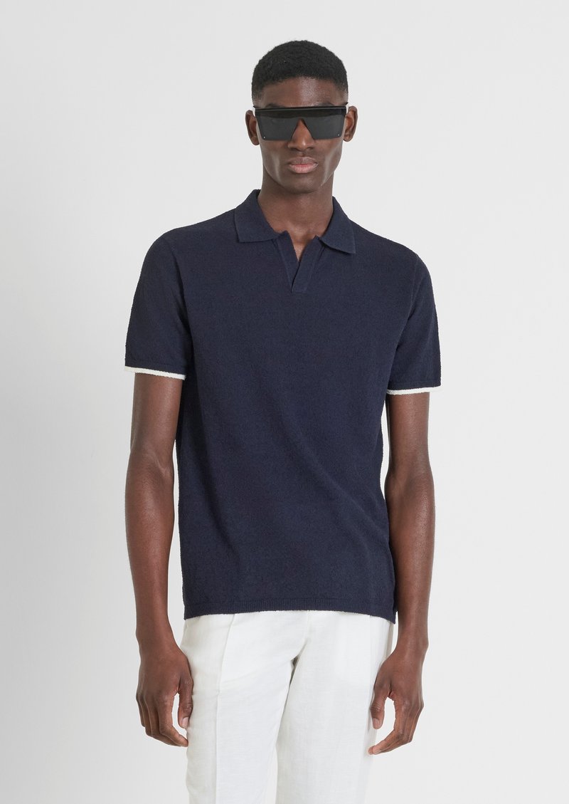 Polo shirt blu navy con bordi bianchi sulle maniche, realizzato in tessuto testurizzato. Indossato con pantaloni chiari, abbinato a occhiali da sole neri.