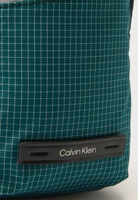 Türkiz színű szövet táska rácsmintával, fekete bőr címkével, amelyen "Calvin Klein" felirat található, valamint texturált felülettel a részletek kiemelésére.