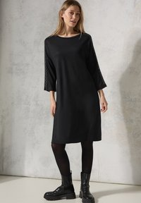 Musta mekko, jossa on 3/4-hihat ja pitsiyksityiskohtia hihoissa. Silkkinen kangas ja väljä malli, yhdistettynä mustiin nilkkabootseihin.