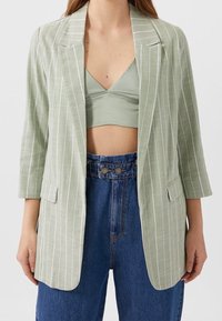 Grüner, gestreifter Blazer mit tailliertem Design, der dreiviertel Ärmel und zwei vordere Taschen aufweist, kombiniert mit einem passenden Oberteil und hochtaillierten Jeans.