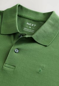 Zöld póló, texturált anyagból készült, gallérral, három gombbal és egy kis hímzett logóval a mellkasnál. Normál fazon.