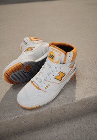 Baskets montantes New Balance 650 blanches et orange posées sur une surface en pierre, mettant en valeur le logo de la marque et les semelles texturées.