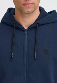 Tamsiai mėlynas zip-up megztinis su kapišonu, su raišteliu, tekstūruota medžiaga ir maža juoda logo ant krūtinės.