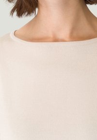 Jasno-beżowy sweter w prążki z dekoltem w łódkę, o miękkiej fakturze i subtelnych detalach przeszyć przy kołnierzu.