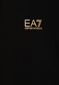 O tecido de algodão preto apresenta um logotipo em destaque dourado com a inscrição "EA7 EMPORIO ARMANI". O logotipo possui uma tipografia moderna com linhas horizontais.