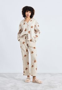 Beige pysjamasett med teddybjørn-mønster, som har en krage med lange ermer og joggebukser med snøring. Myk, teksturert stoff; kombinert med beige tøfler.