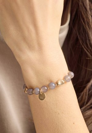 Handgelenk mit einem Perlenarmband mit halbtransparenten grauen Perlen, goldenen Akzenten und einem kleinen goldenen Anhänger mit eingravierten Buchstaben.