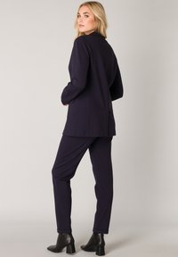Marineblauwe two-piece outfit met een op maat gemaakt blazer en bijpassende broek, vervaardigd uit soepel materiaal. Laarzen met een blokhak maken de look compleet.
