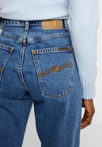 Blå denimjeans med hög midja, kontrasterande orange sömmar och en läderlapp på bakre midjan, med två bakfickor.