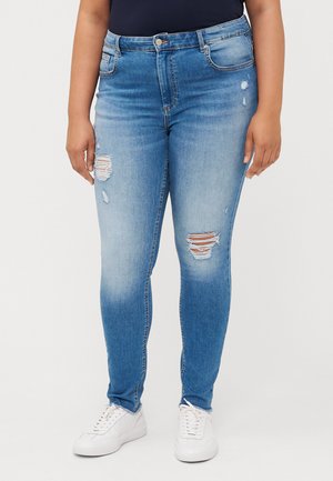 Una persona está de pie vistiendo unos pantalones ajustados rasgados de un azul claro y zapatillas blancas, posando frente a un fondo blanco liso.