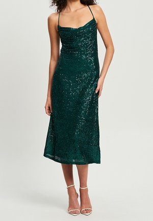 Femme portant une robe midi sans manches, vert foncé pailletée, avec des bretelles fines et des sandales blanches à talons hauts, debout devant un fond uni.