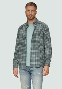 Heren overhemd met knoop in een groen en marineblauw ruitpatroon, vervaardigd uit lichtgewicht stof; gecombineerd met een lichtblauw T-shirt eronder en versleten jeans.