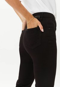 Schwarze Denim-Jeans mit glatter Textur, die ein klassisches Fünf-Taschen-Design, einen mittelhohen Bund und ein rautenförmiges Detail an der Gesäßtasche aufweisen.