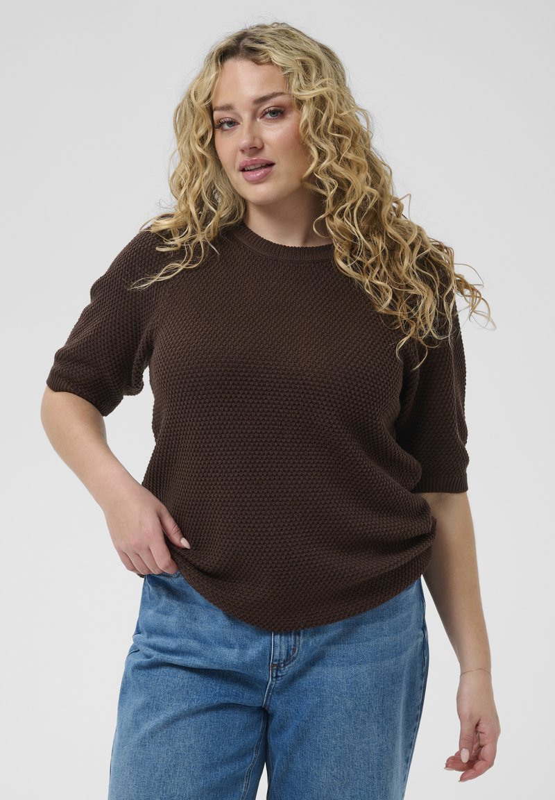 Femme blonde bouclée portant un pull manches courtes texturé marron foncé et un jean bleu clair, debout devant un fond uni.