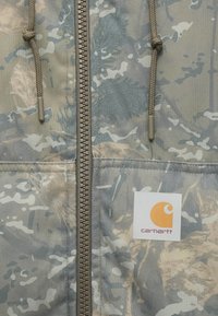 Maskáčová bunda s prackou na zips, v kaki a sivej farbe, s textúrovanou látkou a štítkom s logom Carhartt v pravom dolnom rohu.