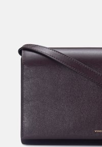 Sac bandoulière en cuir violet foncé avec une sangle fine, une texture lisse et un petit lettrage doré de la marque dans le coin inférieur droit.