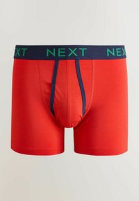 Boxeri roșii pentru bărbați cu talie bleumarin, având text verde „NEXT” și o margine verticală bleumarin în jurul zonei buzunarului frontal.