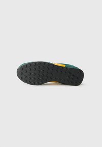 Adidași cu o talpă din cauciuc negru, având un model circular pentru aderență, un superior din piele de căprioară verde și accente galbene pe lateral.