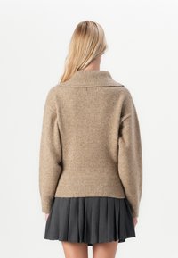 Beige ullblanding genser med høy krage, ribbestrikket nederkant og vide ermer, kombinert med en mørk plisserte skjørt. Teksturert finish er synlig.