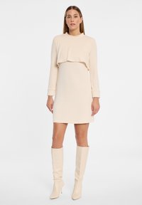 Langärmliges cremefarbenes Kleid mit einer gesteppten Textur, das eine verkürzte obere Schicht aufweist und mit kniehohen cremefarbenen Stiefeln kombiniert wird.