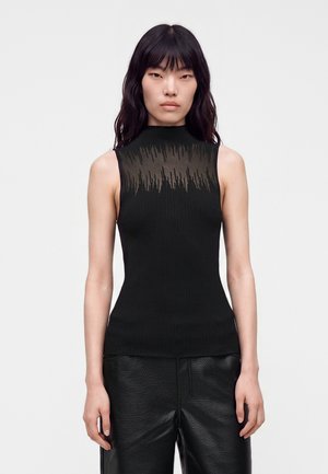 Donna con capelli lunghi e scuri che indossa una maglia nera senza maniche con un giogo trasparente a fantasia e pantaloni neri texturizzati, in piedi su uno sfondo semplice.
