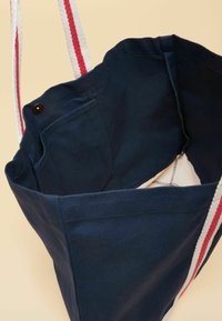 Tumesinine kandekott valgete ja punaste triibuliste sangadega, näidates sisemist taskut ja neetidega kinnitust, beeži tausta ees.