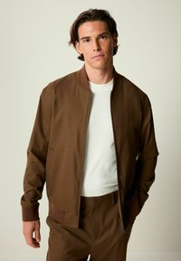 Braune Bikerjacke mit einer einheitlichen Textur, gerippten Bündchen und einem Reißverschluss. Getragen über einem weißen Hemd, kombiniert mit passenden braunen Hosen.