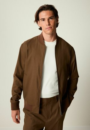 Brown bomber jacket with a solid texture, ribbed cuffs, and a zip front. Worn over a white shirt, paired with matching brown trousers.