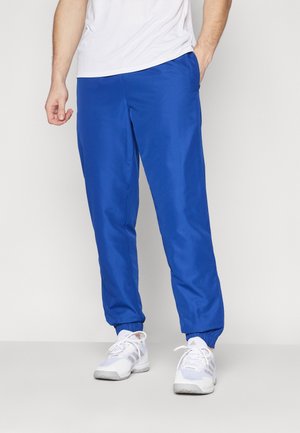 Man draagt felblauwe joggingbroek met elastische boorden en witte sportschoenen, staat met één hand in de zak.