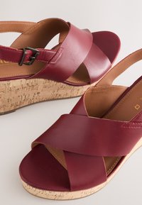 Rote Keilsandalen aus Leder mit Korksohle, mit Kreuzriemen-Design und verstellbarer Schnalle. Glatte Textur mit kontrastierenden Akzenten.