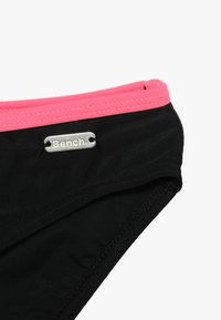 Svart svømmebukse med en rosa elastisk midjebånd, med et sølvfarget metallmerke med påskriften "Bench." Glatt stofftekstur.