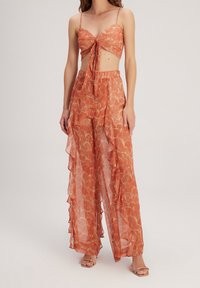 Conjunto de dos piezas que incluye un top corto con lazo en el frente y pantalones anchos con volantes. Fabricado en una tela ligera y transparente con un patrón de conchas en naranja y blanco.