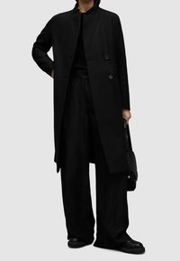 Czarny wełniany płaszcz z wysokim kołnierzem i podwójnym zapięciem na guziki, w połączeniu z luźnymi czarnymi spodniami i czarnymi butami na masywnej podeszwie. Minimalistyczny design.