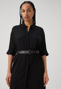 Schwarzes Hemdkleid mit kurzen Ärmeln, das eine taillierte Taille aufweist, die durch einen breiten schwarzen Gürtel mit runden Ausschnitten und einer Metallschnalle betont wird.
