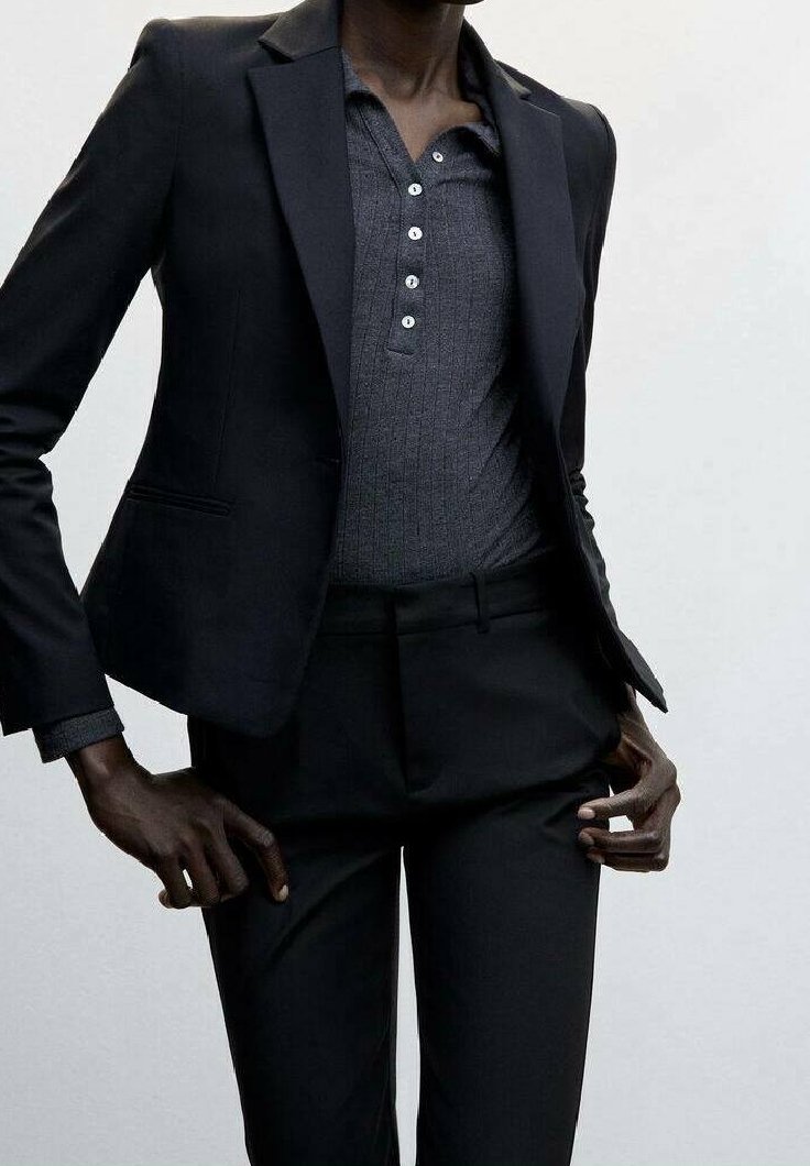 Personne portant un blazer noir cintré sur une chemise gris foncé boutonnée et un pantalon noir, sur un fond clair uni.