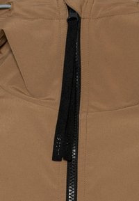 Brun jacka med en central svart dragkedja, texturerad tyg och en dragkedjehandtag med logotyp. Tydlig sömnad och synliga fickkanter.