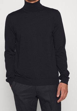 Homme portant un pull noir moulant à col roulé avec manches longues et pantalon gris foncé à fines rayures, debout avec une main dans la poche.