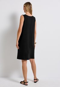 Robe noire sans manches avec une coupe décontractée, texture lisse et encolure arrondie. Associée à des sandales noires à lanières, vue de dos.
