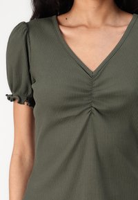 Top a coste verde oliva con scollatura a V, centro arricciato e maniche puff con polsini arricciati, realizzato in un tessuto morbido ed elastico.