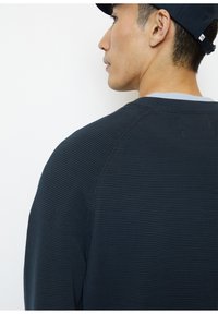 Marineblauer Strickpullover mit strukturiertem Stoff, geripptem Kragen und Raglanärmeln. Die Rückansicht hebt die lässige Passform und die Details des Kleidungsstücks hervor.