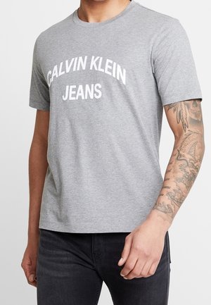 Mann trägt ein graues Calvin Klein Jeans T-Shirt und schwarze Jeans, zeigt einen detaillierten Tattoärmel auf seinem rechten Arm.