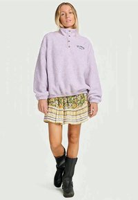 Pullover in pile viola chiaro con colletto alto e tre bottoni dorati. Abbinato a una gonna a fantasia floreale e stivali neri fino al ginocchio.