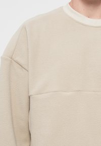 Pull en polaire beige avec une surface texturée, présentant un col rond et un détail de couture horizontal sur la poitrine. Tissu doux.