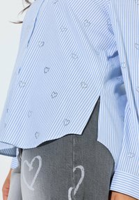 Blau-weiß gestreiftes Hemd mit kleinen Strassherzmuster, kombiniert mit grauer Hose mit weißen Herzmotiven.