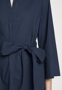 Robe bleu marine à manches longues avec une large ceinture en tissu nouée en nœud à la taille, présentée du buste jusqu'à la mi-cuisse.
